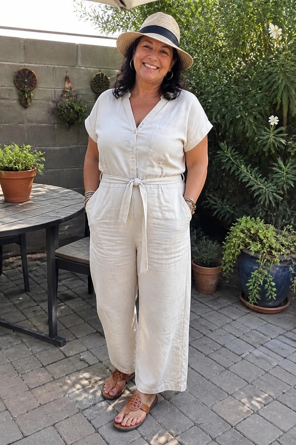 Smiling woman in cream jumpsuit and straw hat stands on brick patio.