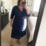 Woman in navy wrap dress with brown belt taking mirror selfie in bedroom