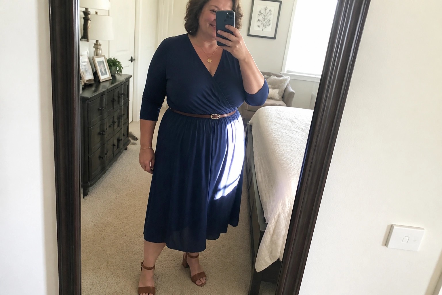 Woman in navy wrap dress with brown belt taking mirror selfie in bedroom