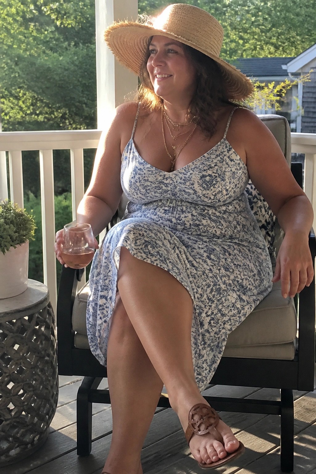 Woman in blue floral dress and straw hat sits on porch holding wine glass