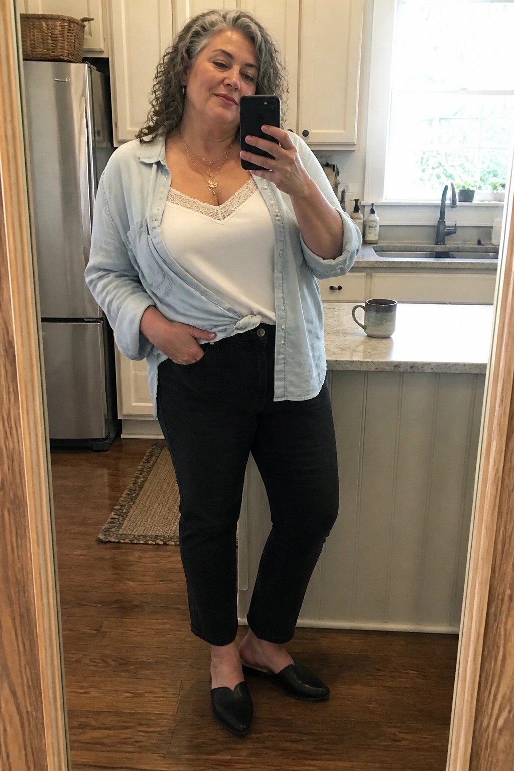 Gray-haired woman in light blue tied shirt and black jeans taking kitchen mirror selfie.