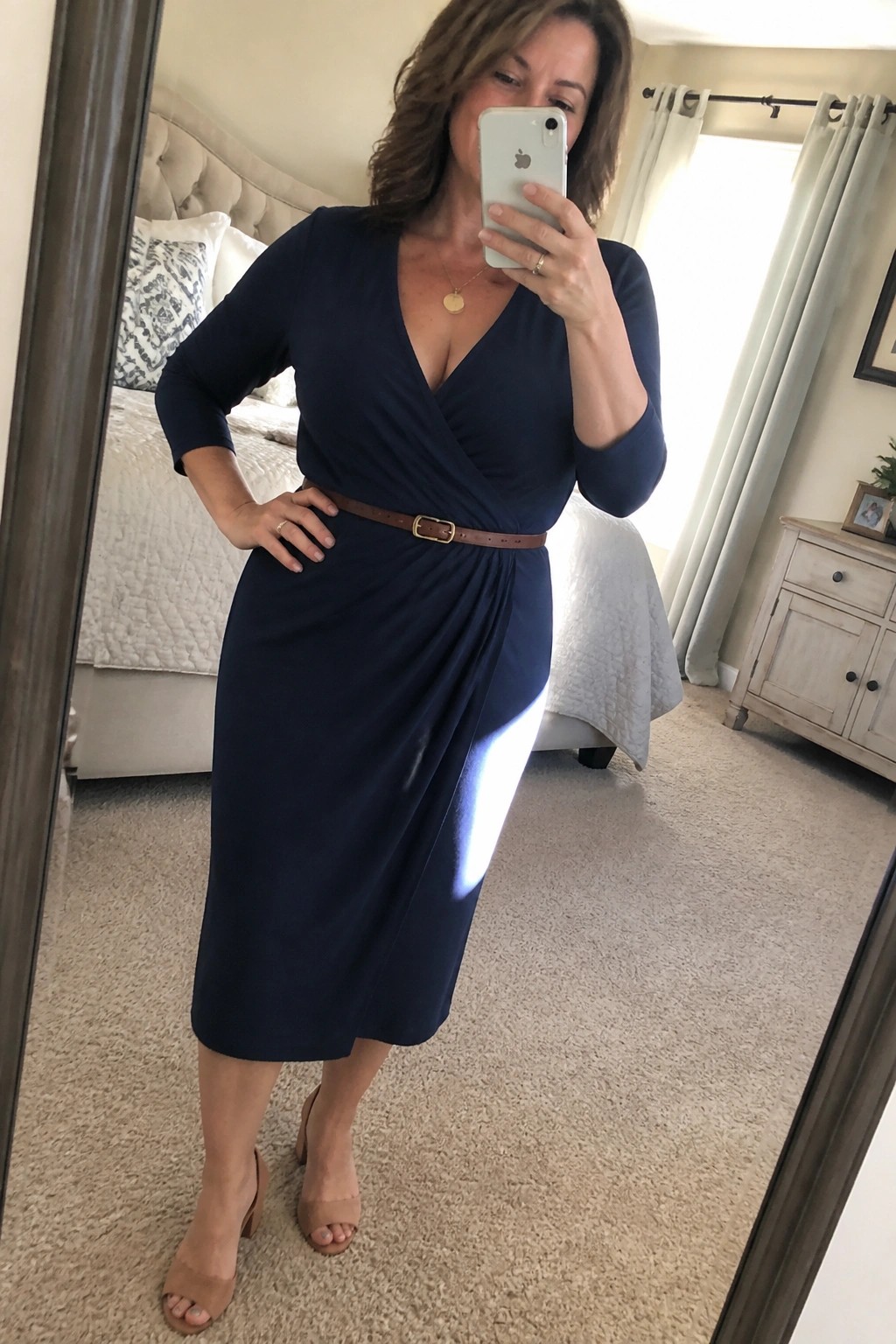 Woman in navy wrap dress with brown belt taking mirror selfie in bedroom