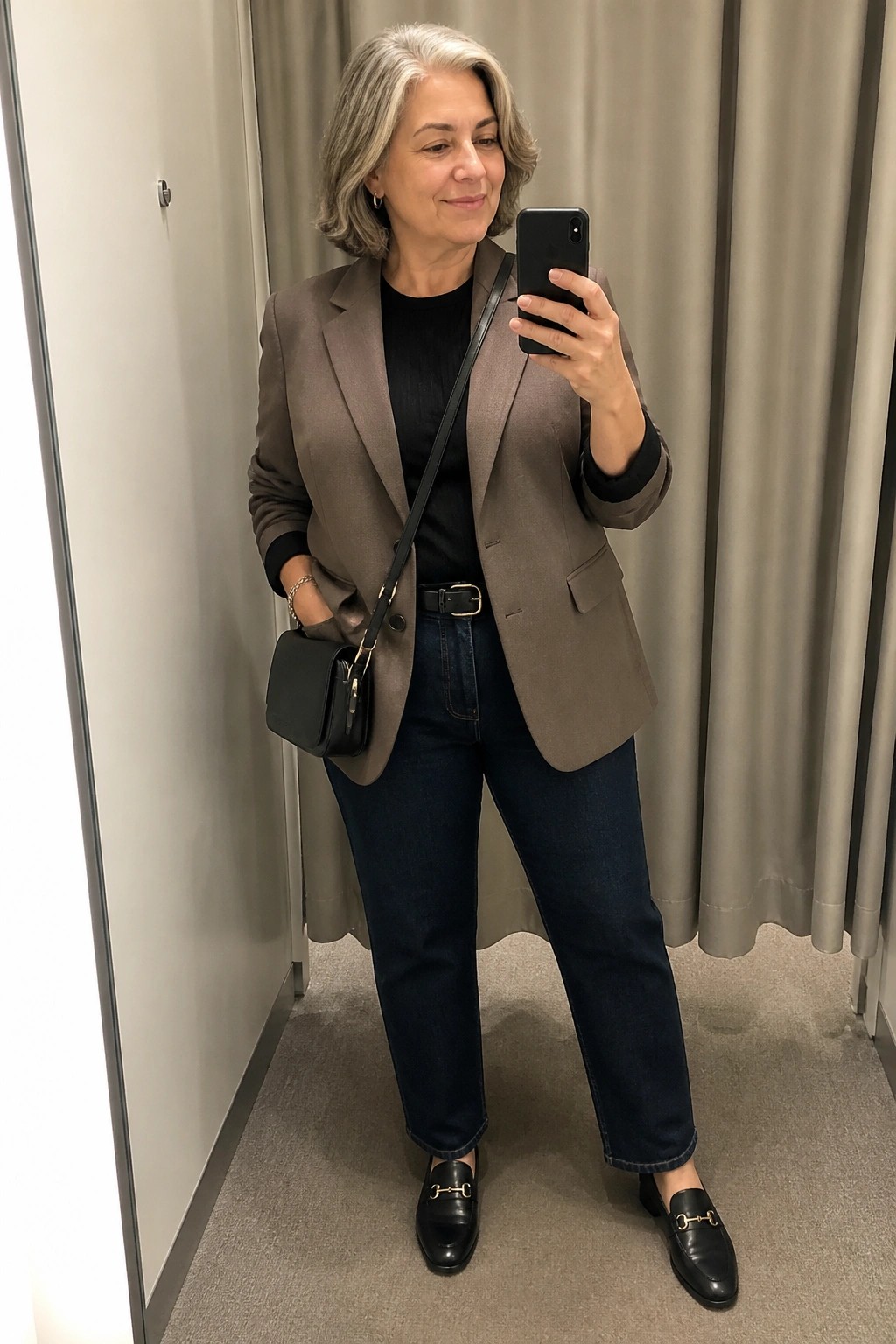 Gray-haired woman in taupe blazer and jeans taking mirror selfie in fitting room.