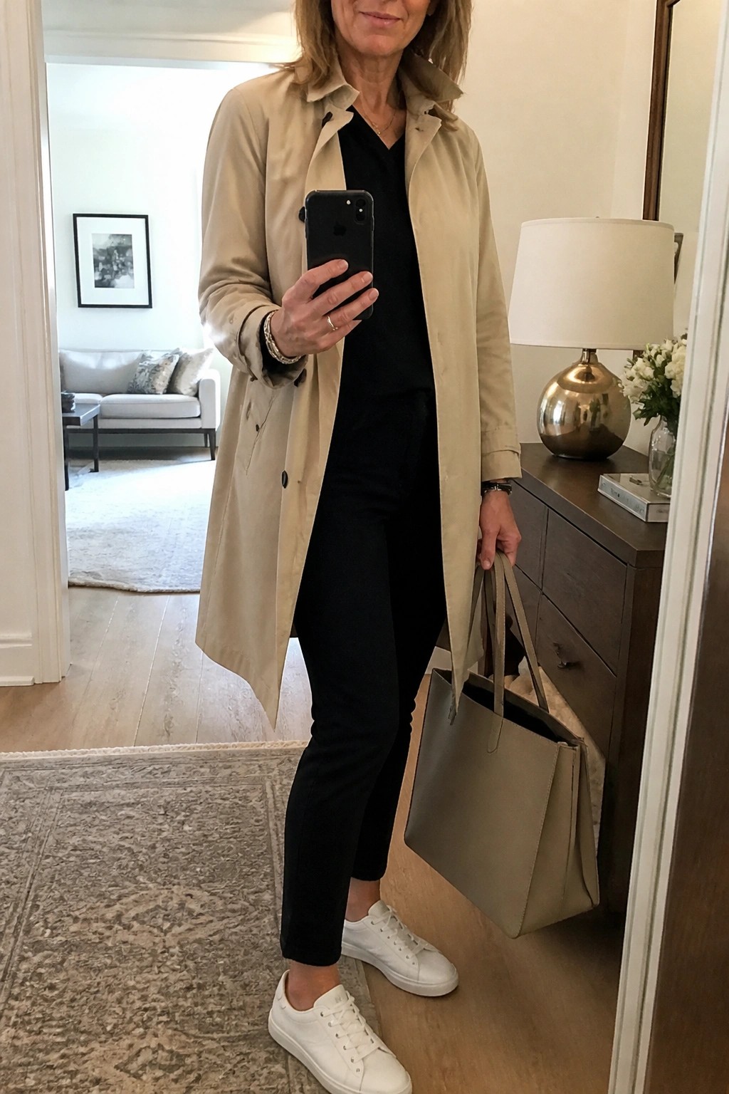 Woman in beige trench coat, black top and pants, white sneakers, holding tote and phone