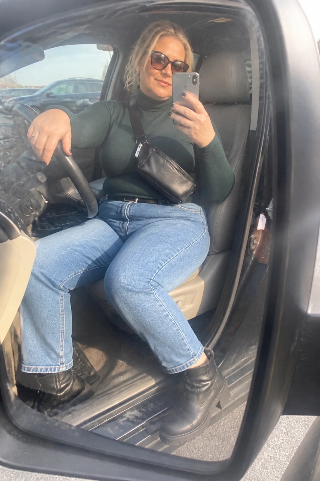 Blonde woman in green turtleneck and jeans taking car selfie with phone