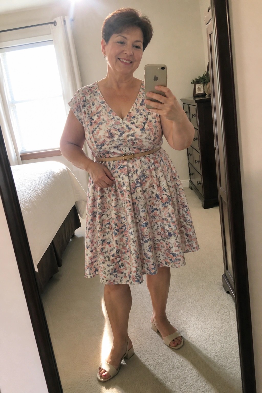 Woman in floral dress with belt taking bedroom mirror selfie in beige heels