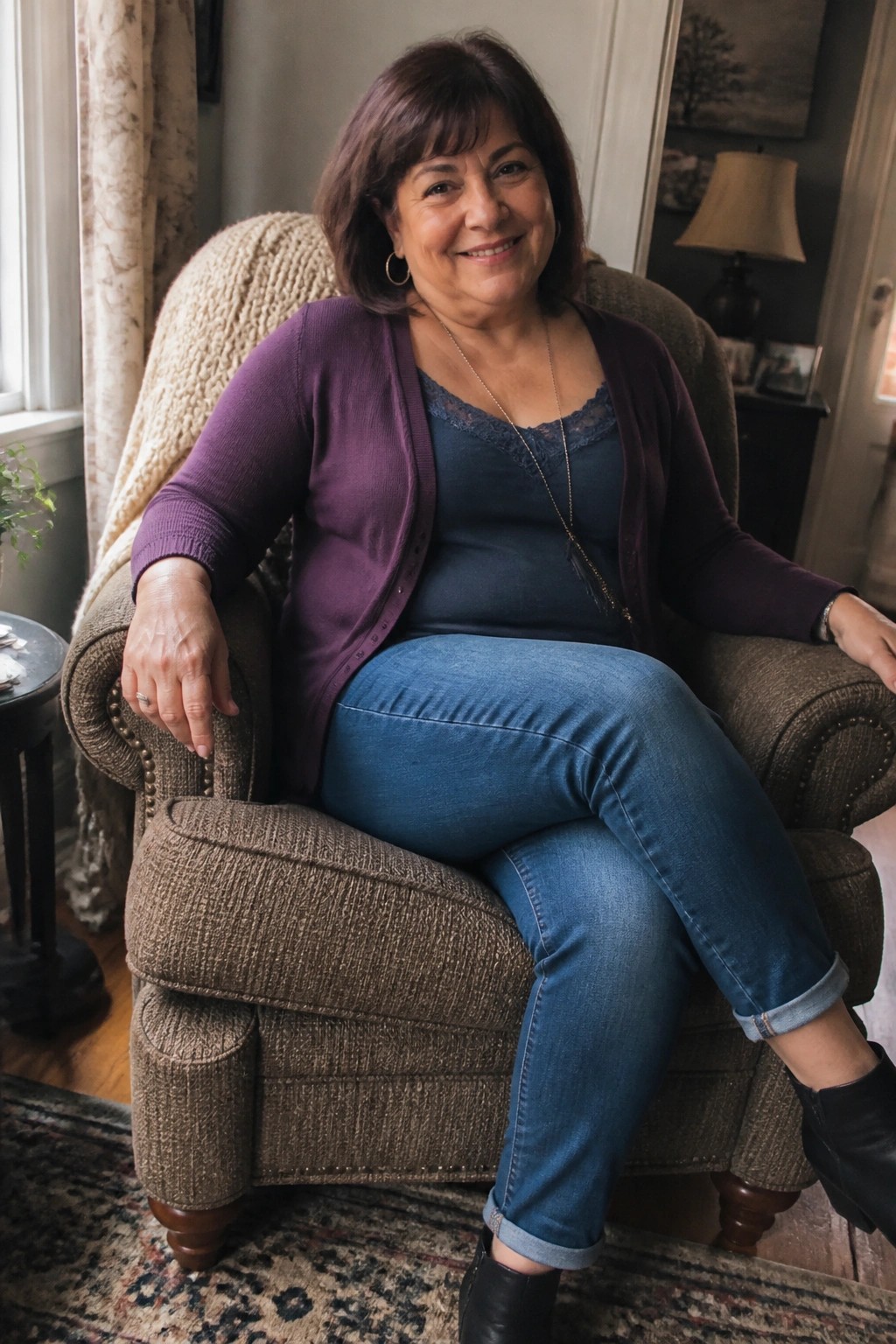 Woman over 50 wearing purple cardigan over blue lace cami with cuffed blue jeans and black ankle boots plus long necklace seated in brown armchair