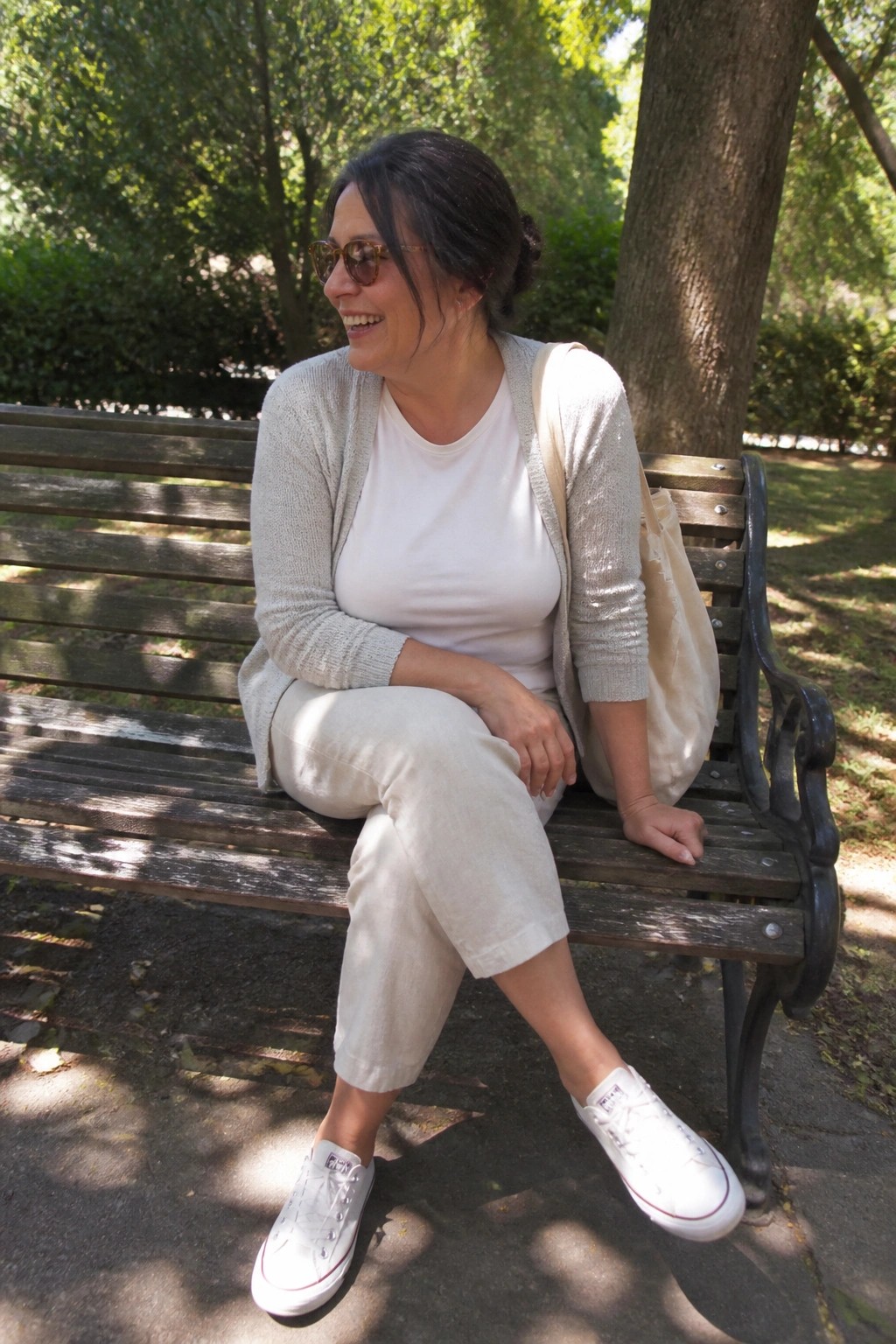 Smiling woman in beige cardigan and white sneakers on sunny park bench
