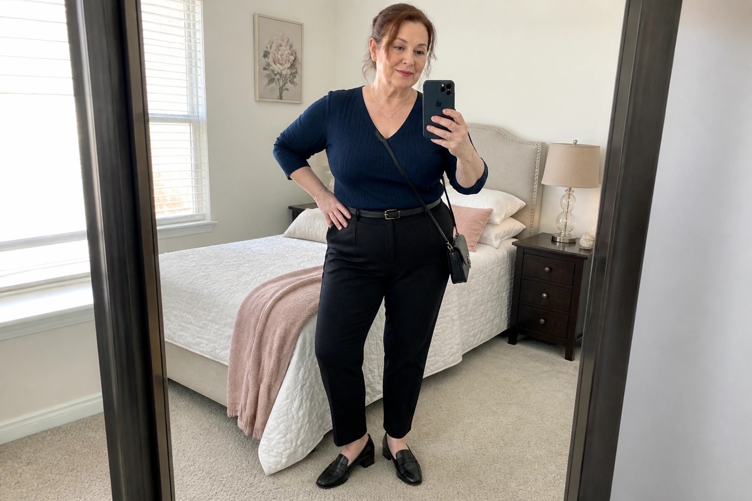 Woman in navy sweater and black pants taking mirror selfie in bedroom