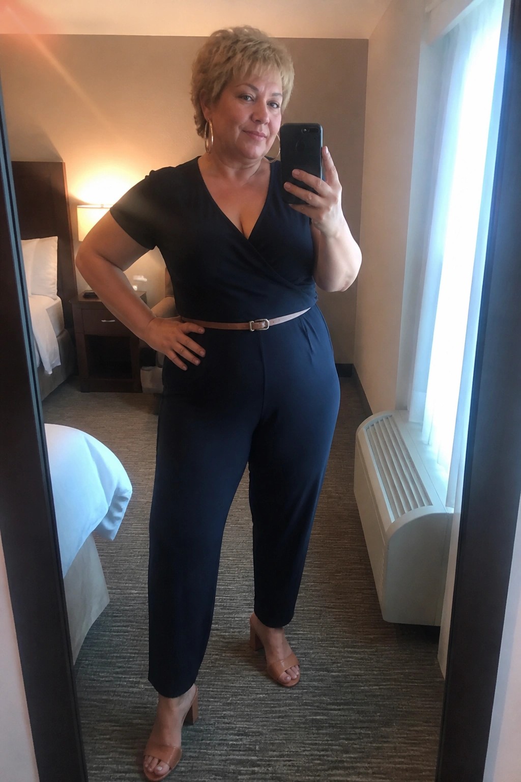 Woman in black jumpsuit taking mirror selfie in hotel room with brown belt and heels