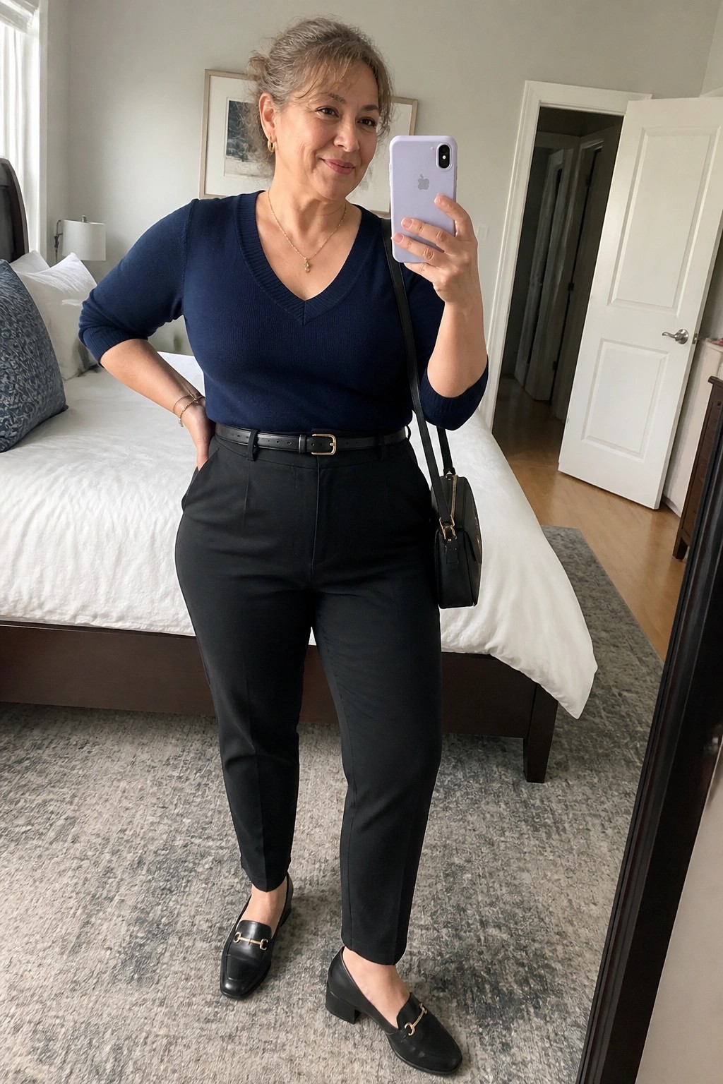 Woman in navy sweater and black pants taking mirror selfie in bedroom