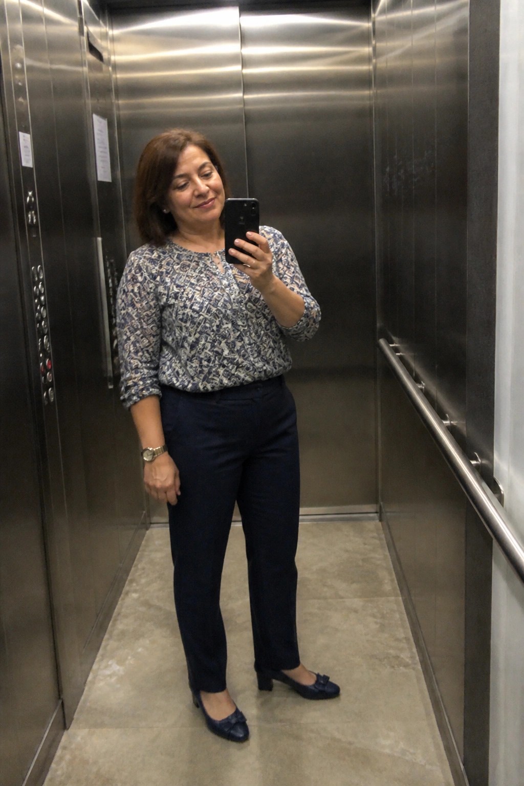 Woman in patterned blouse taking selfie inside stainless steel elevator.