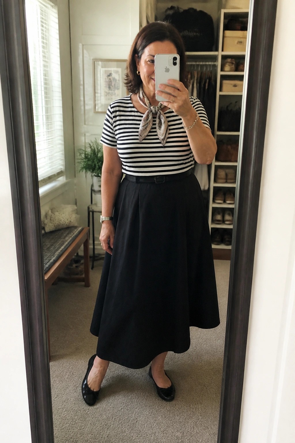 A woman wearing a black and white striped short sleeve top tied with a patterned scarf at the neck, a black midi A-line skirt with a thin belt, and black ballet flats while taking a mirror selfie in a closet area.