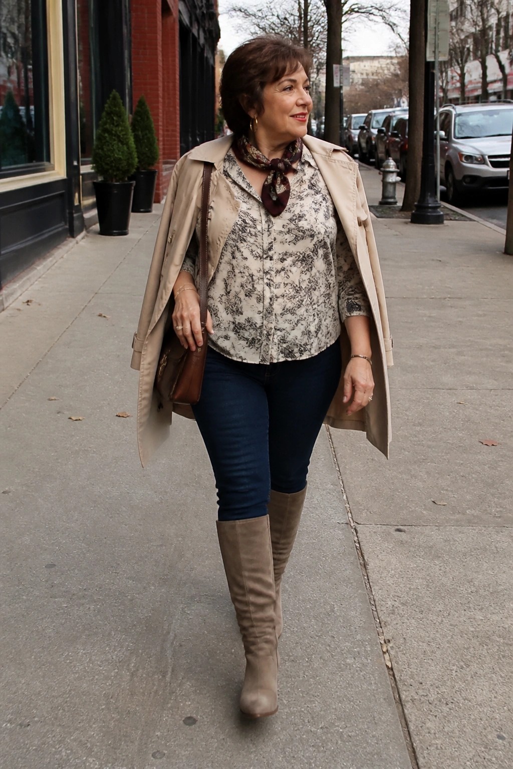 Woman in beige trench coat, floral blouse, jeans, and boots on sidewalk