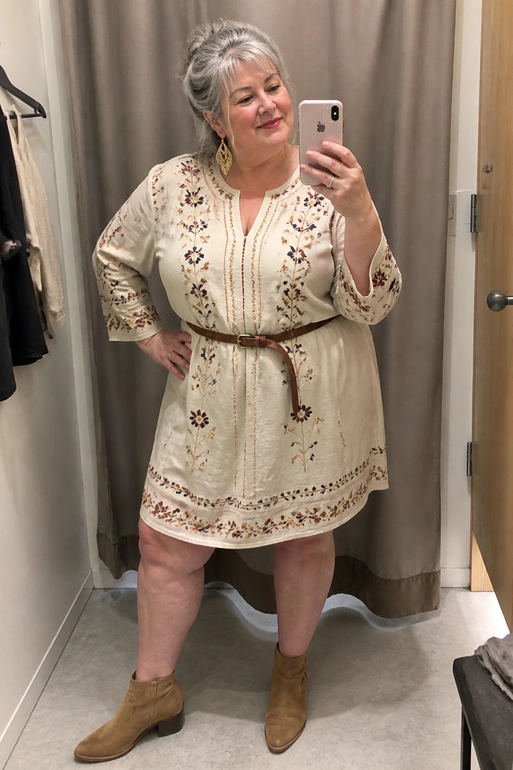 Woman with gray hair in cream embroidered dress and brown boots takes mirror selfie.