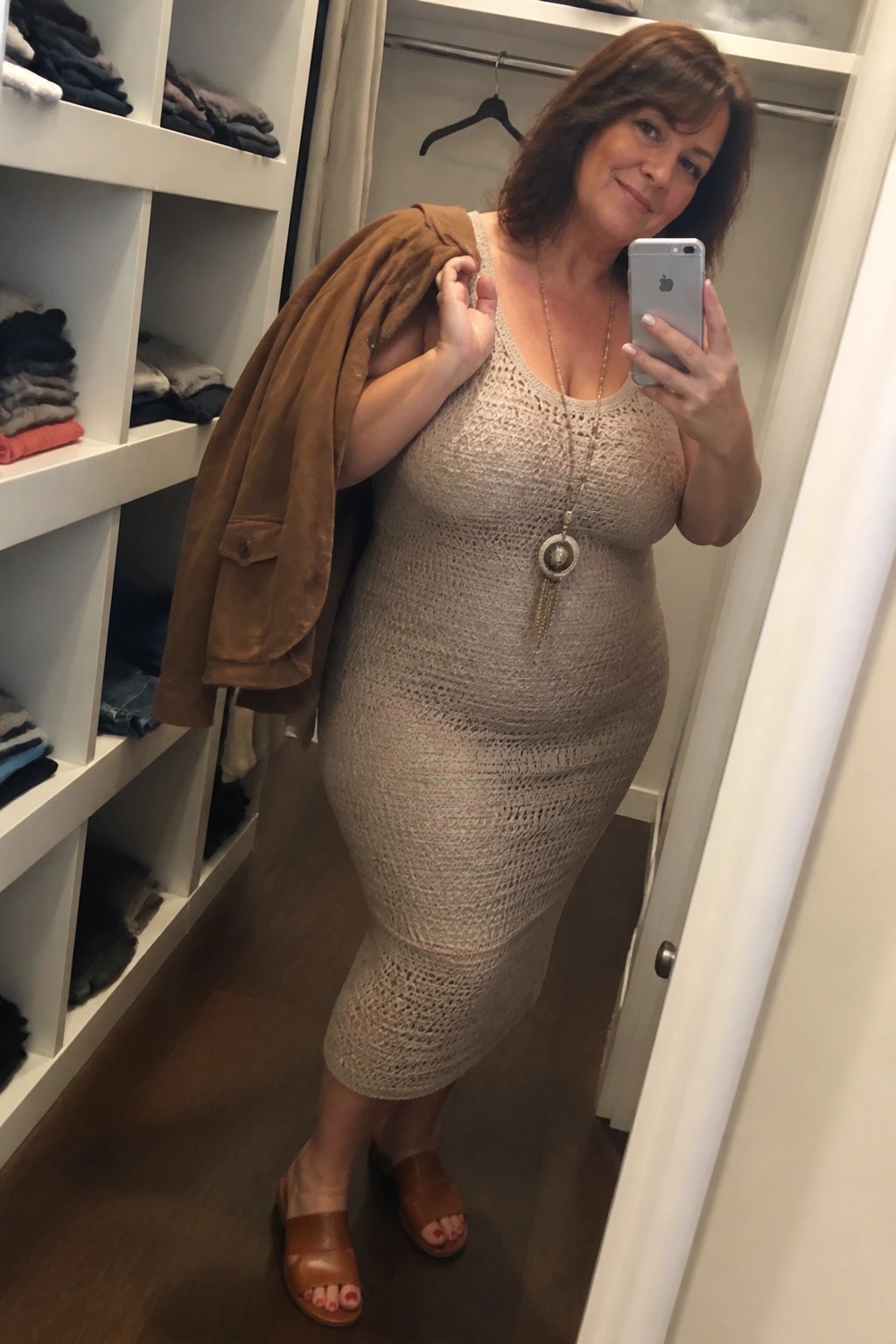 Woman in a beige crochet midi dress holding a brown suede jacket over her shoulder paired with a long pendant necklace and brown slide sandals.