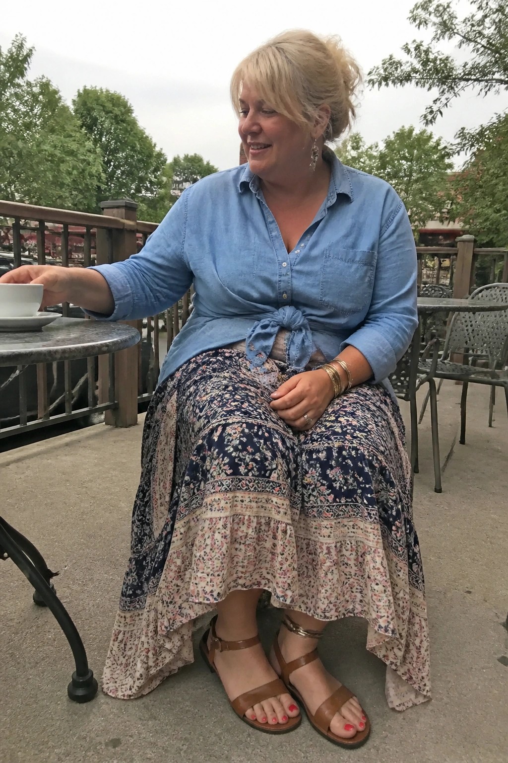 Woman seated outdoors wearing a light blue denim shirt tied in a front knot over a tiered navy and pink floral maxi skirt with ruffles, paired with brown strappy sandals and gold bangles, holding a white coffee cup.