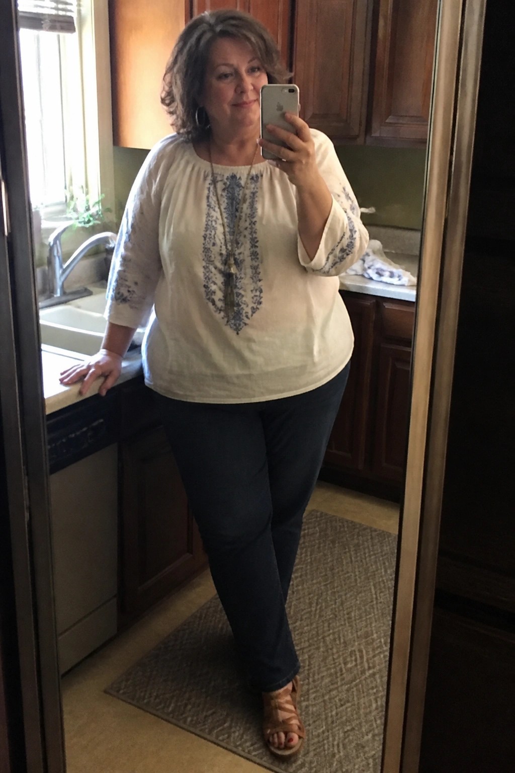Woman wearing a cream embroidered boho blouse with dark jeans and brown strappy sandals while taking a mirror selfie