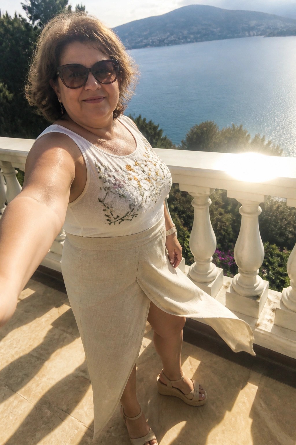 Woman in embroidered white top and beige skirt on sunny seaside balcony overlooking sea