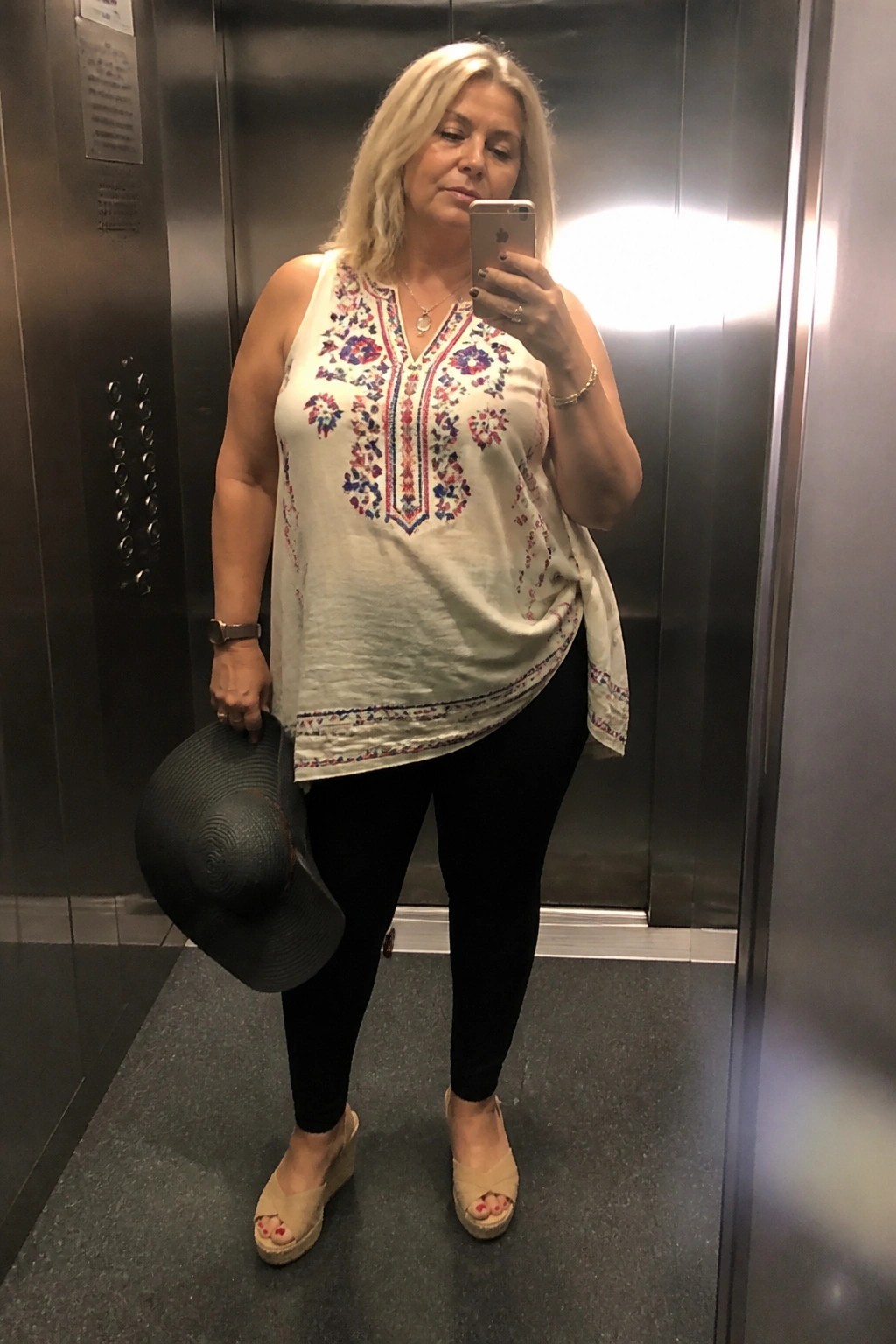 Blonde woman in embroidered white top takes mirror selfie in elevator holding black hat.
