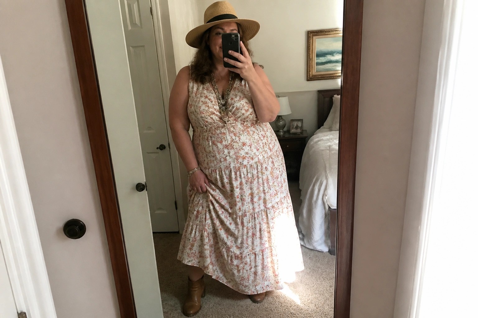 Woman in floral tiered dress, straw hat, and boots taking bedroom mirror selfie