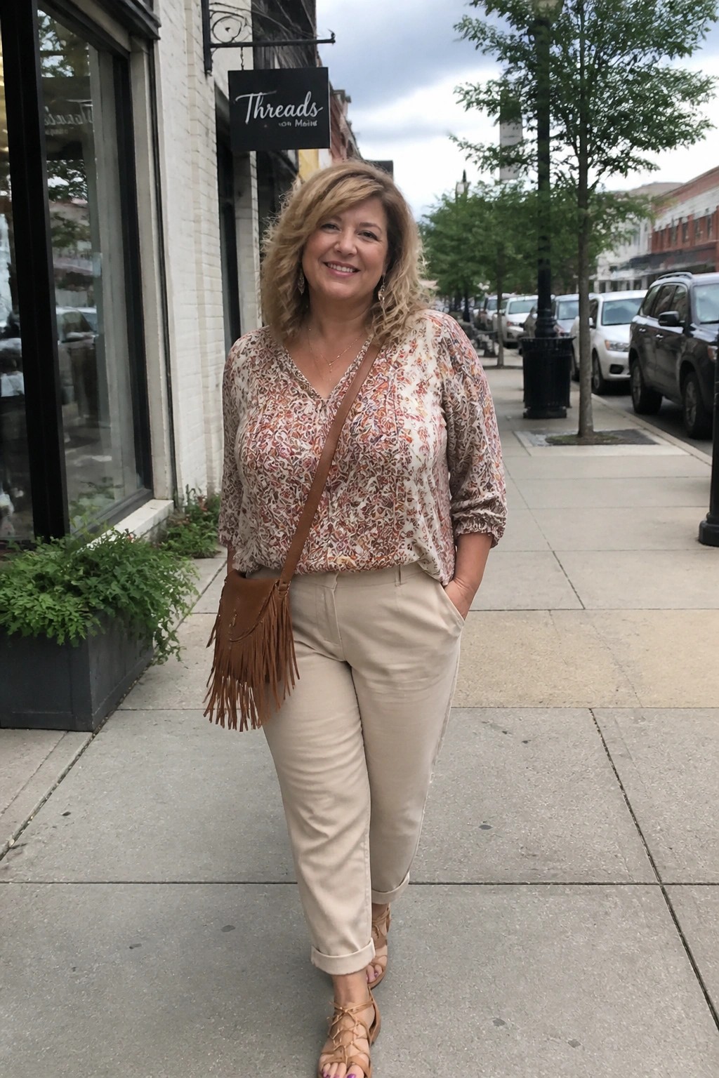 Smiling woman in floral blouse and beige pants with fringed bag on sidewalk