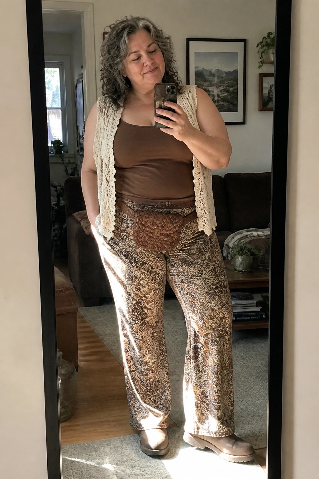 Woman with curly gray hair in brown top, lace vest, and patterned pants taking mirror selfie