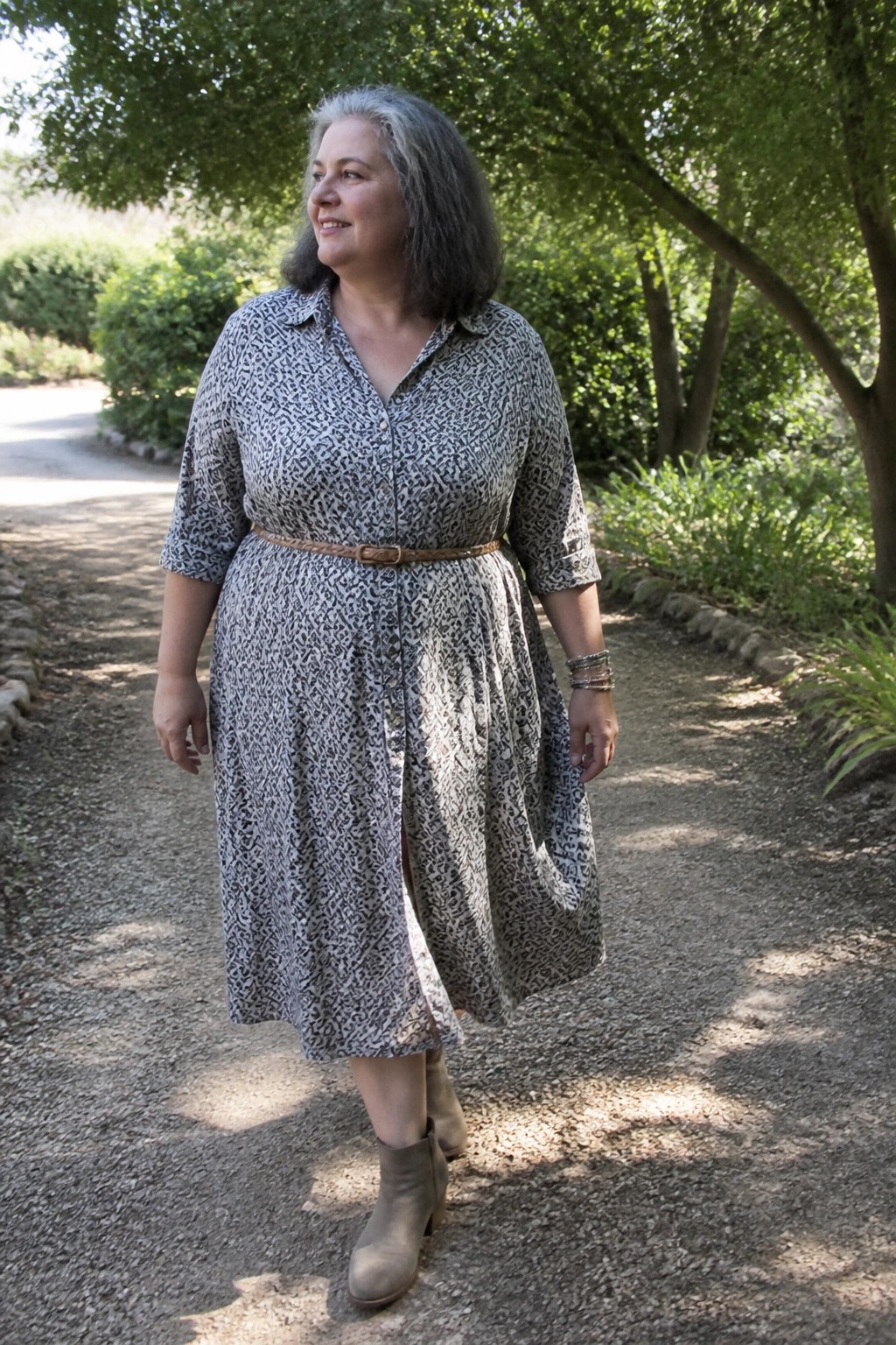 Woman with gray hair in patterned belted dress walks on sunny garden path