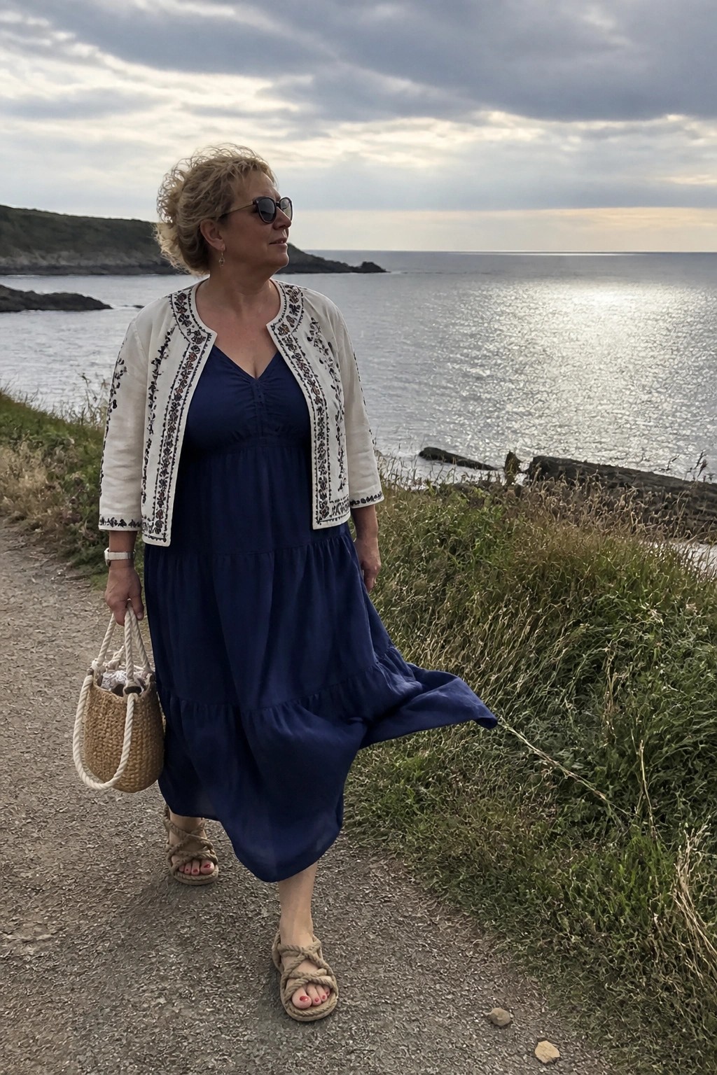 Woman in blue tiered dress and embroidered white jacket holding straw bag by sea