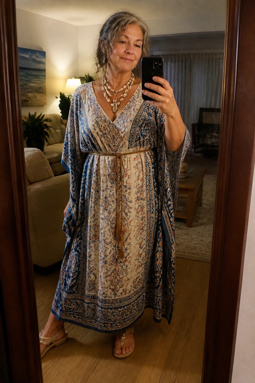Gray-haired woman in blue floral kaftan takes mirror selfie in living room.