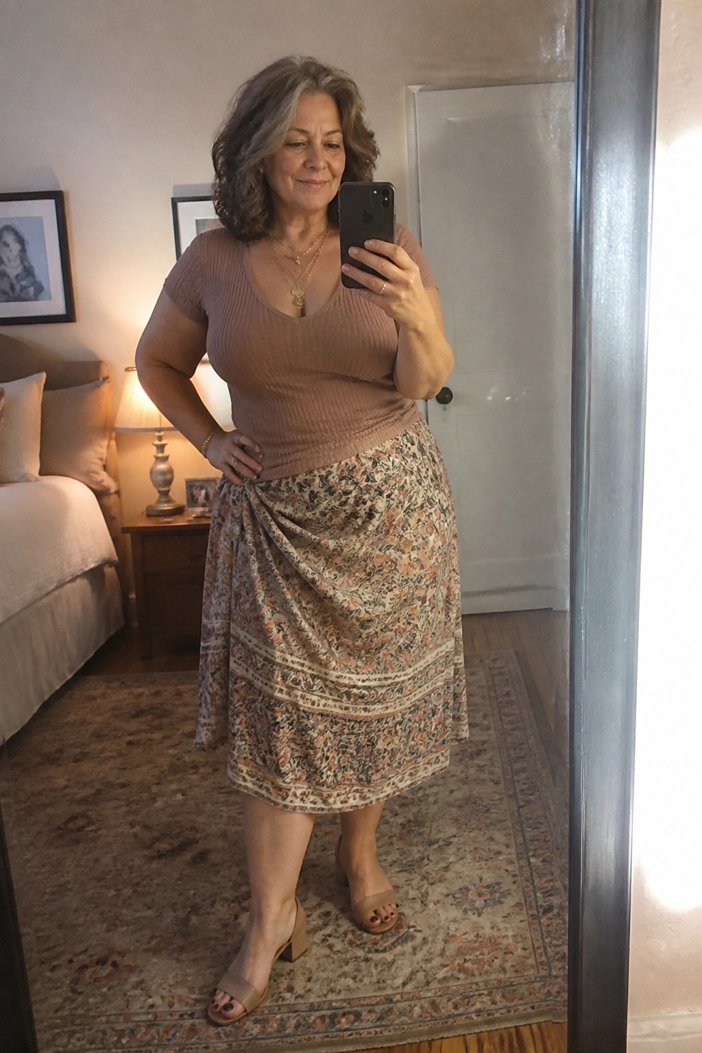 Plus size woman over 50 wearing a dusty rose ribbed short sleeve top tucked into a floral print midi skirt with wrap tie and striped borders, beige block heel sandals, and layered gold necklaces in a bedroom mirror selfie.