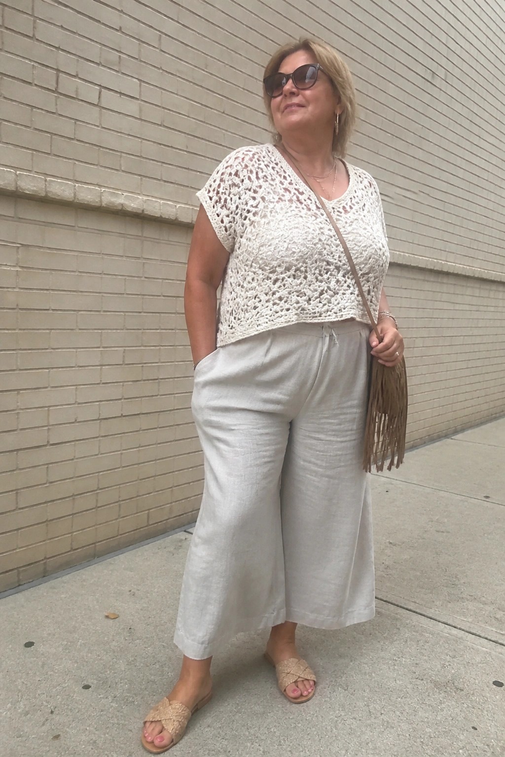Woman in white crochet top and beige wide pants with fringed bag by brick wall
