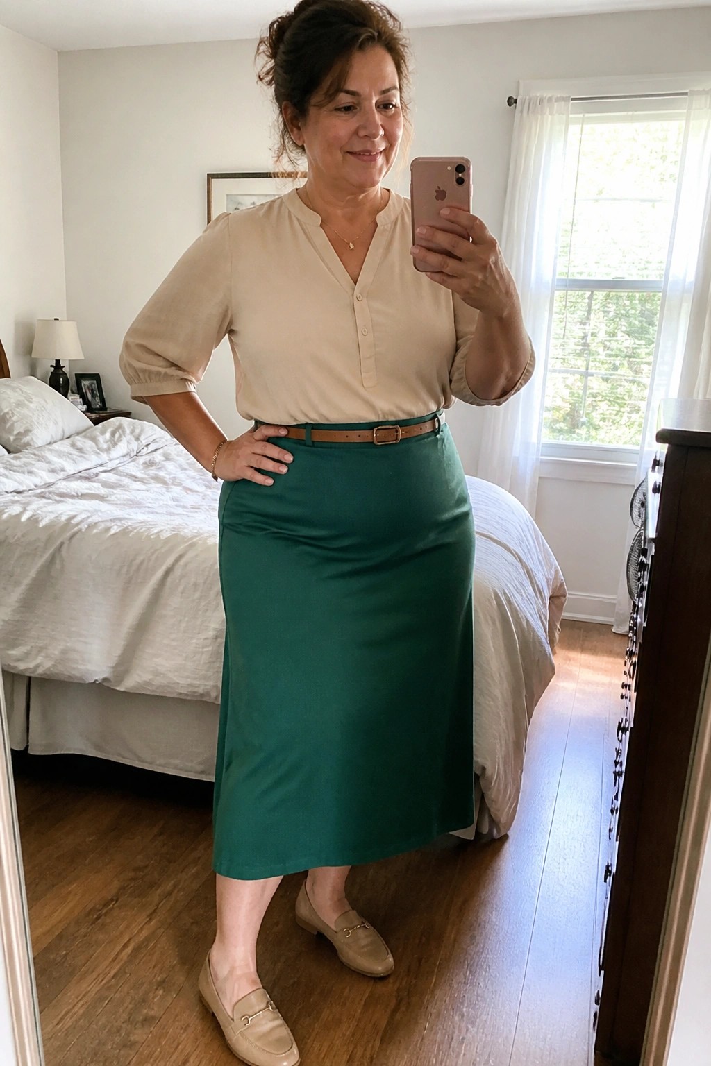 Woman in beige blouse and green midi skirt taking mirror selfie in bedroom.