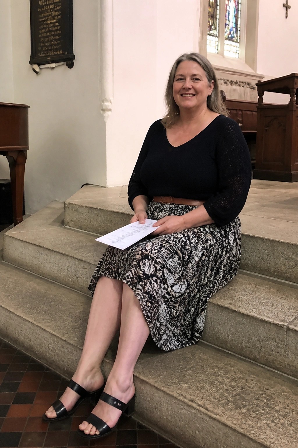 Woman in black top and patterned skirt sits on church steps holding paper