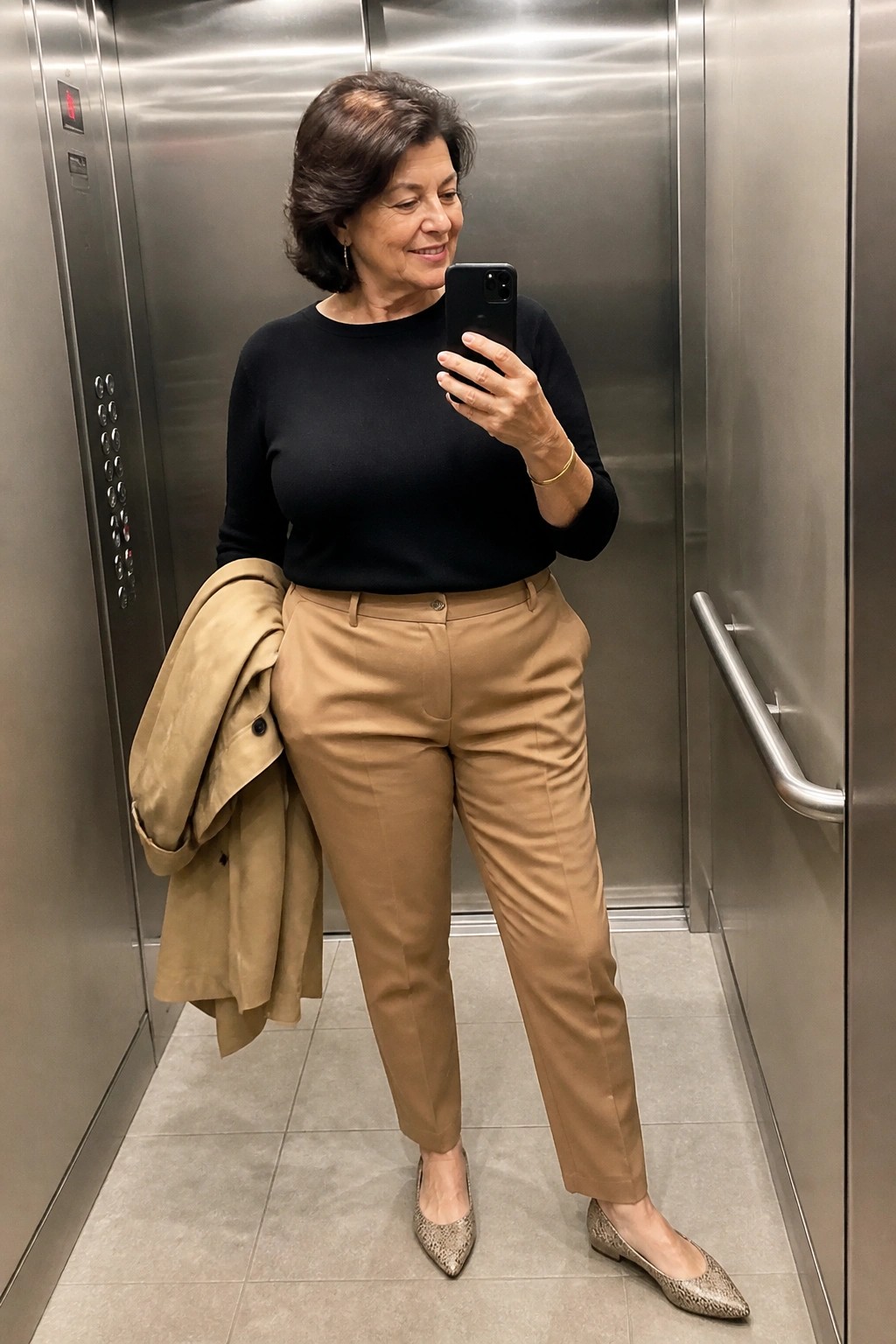 Woman in black top and tan pants takes selfie in elevator holding coat
