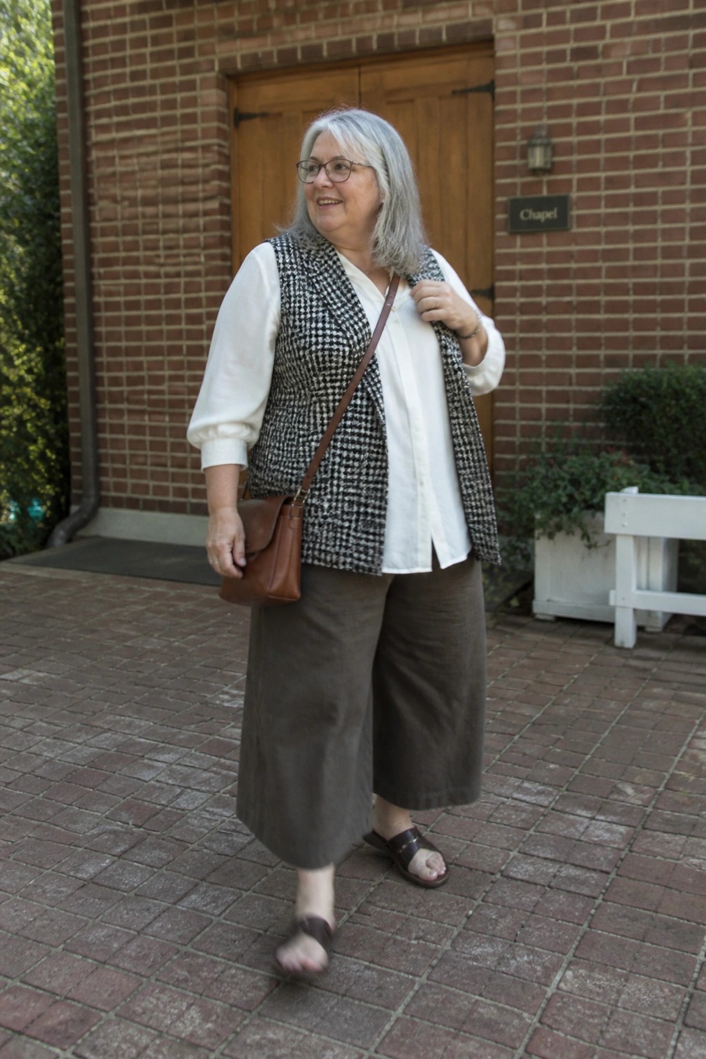 Woman in houndstooth vest and brown wide-leg pants stands before brick chapel.