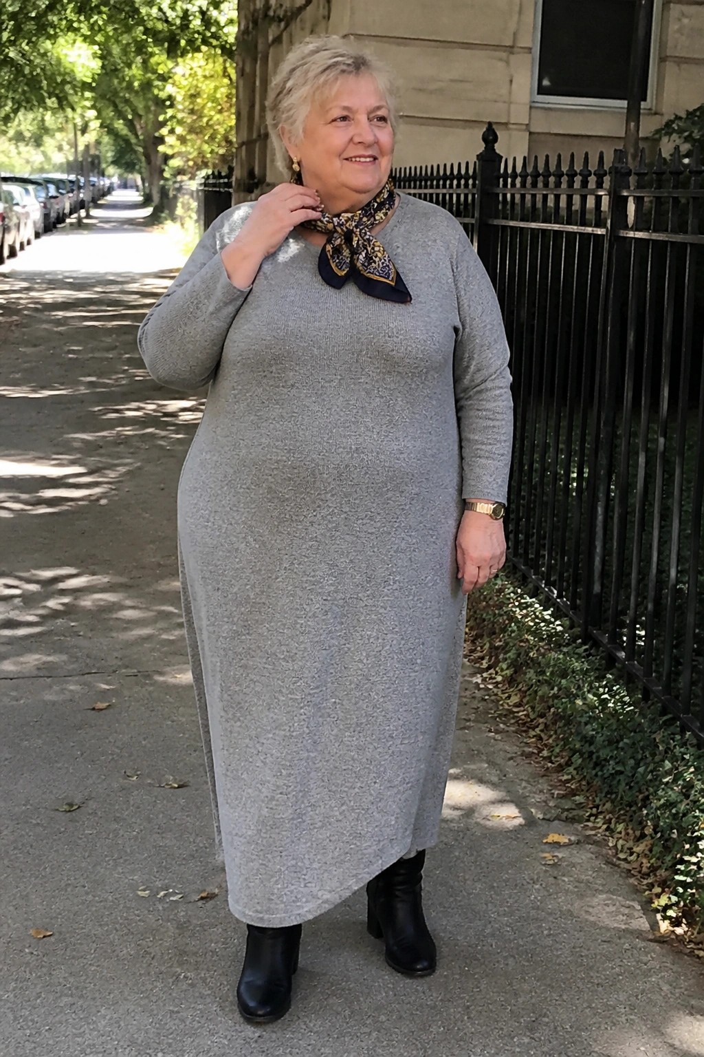 Smiling older woman in long gray knit dress with scarf and boots on sidewalk