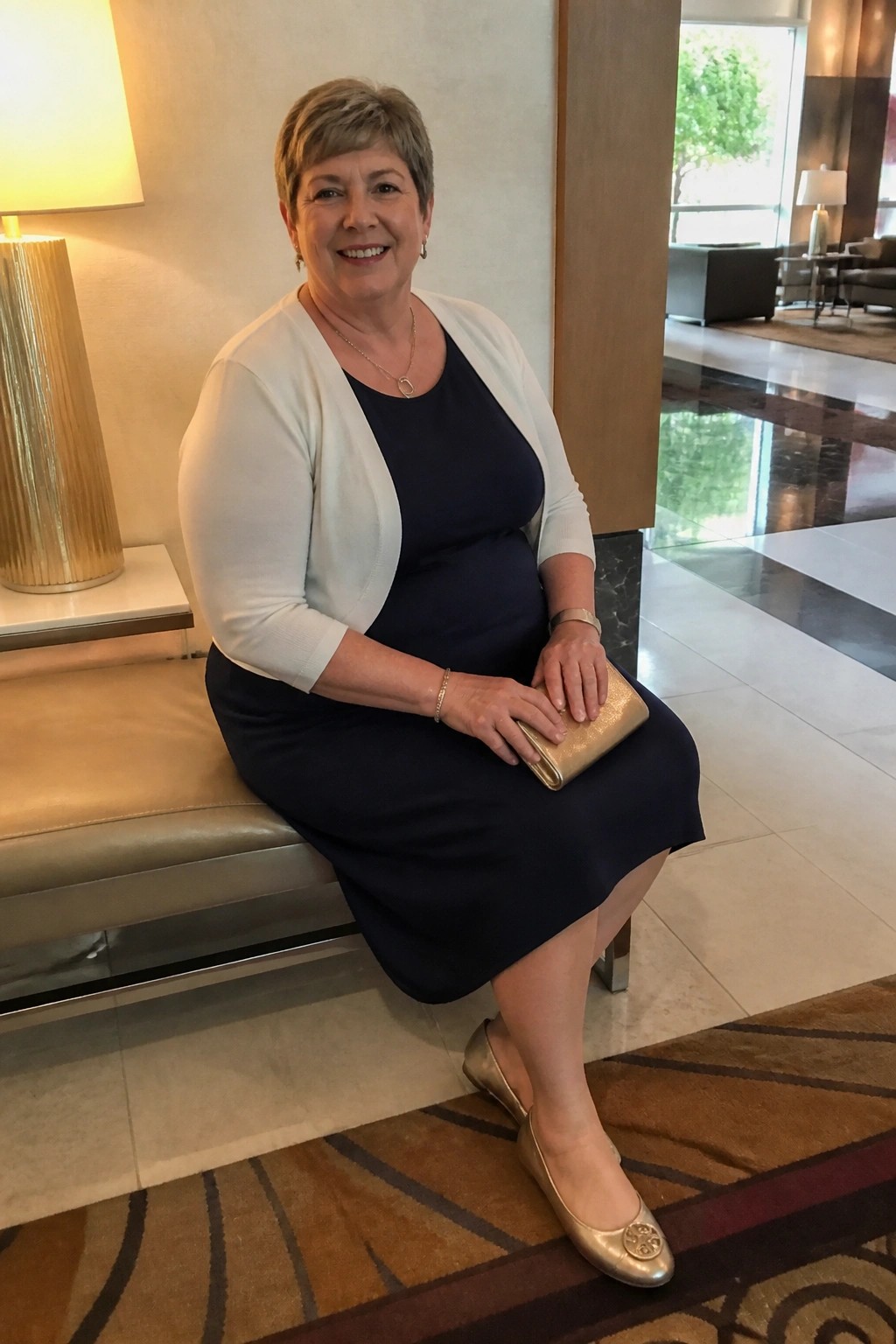 Smiling woman in navy dress and white cardigan with gold clutch seated in lobby