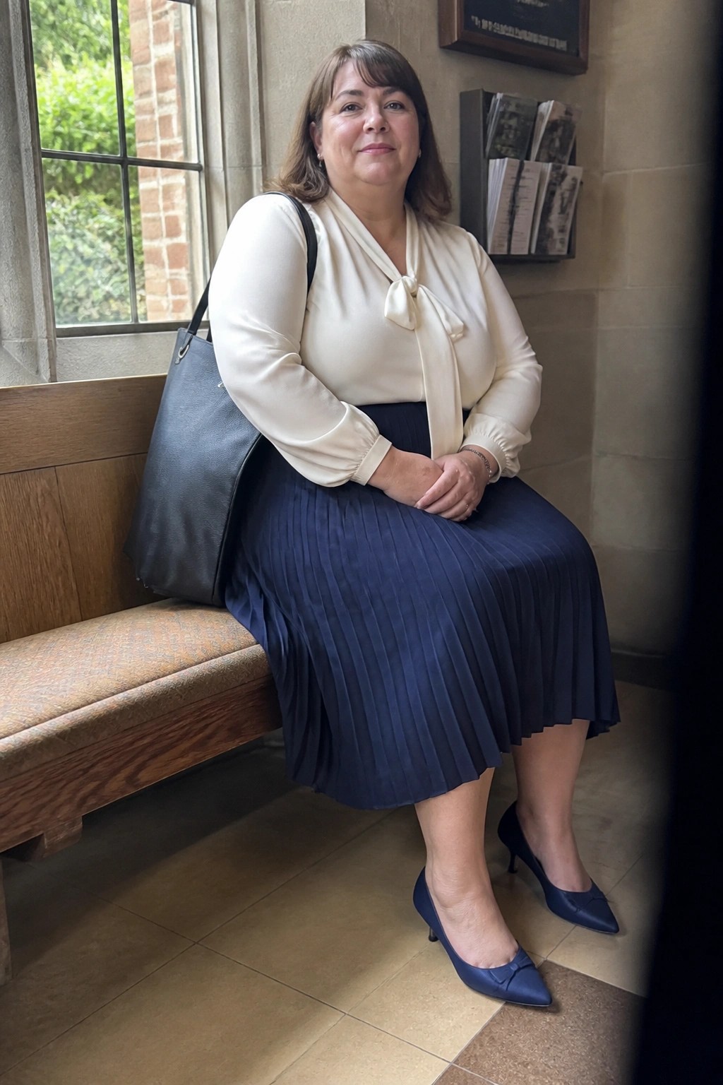 Woman in cream bow blouse and navy pleated skirt seated on bench with black bag
