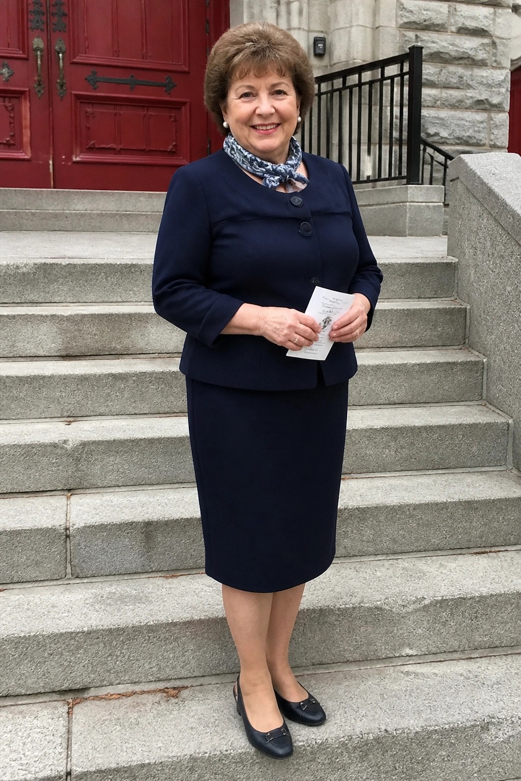 Smiling woman in navy blue suit and scarf holding paper on stone steps