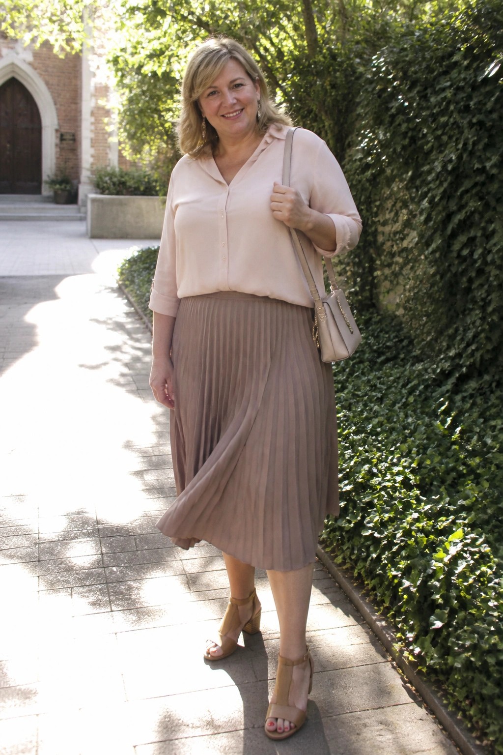 Smiling woman in light pink blouse and taupe pleated skirt holding handbag on path
