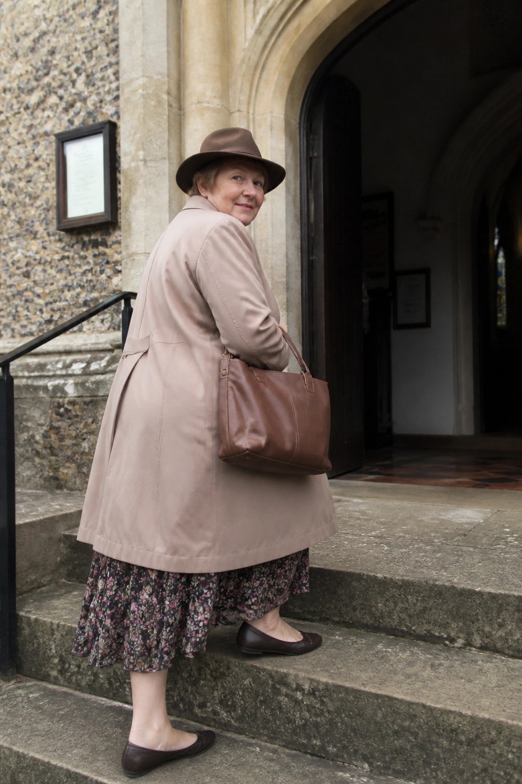 Elderly woman in beige coat and brown hat with leather bag on stone steps.