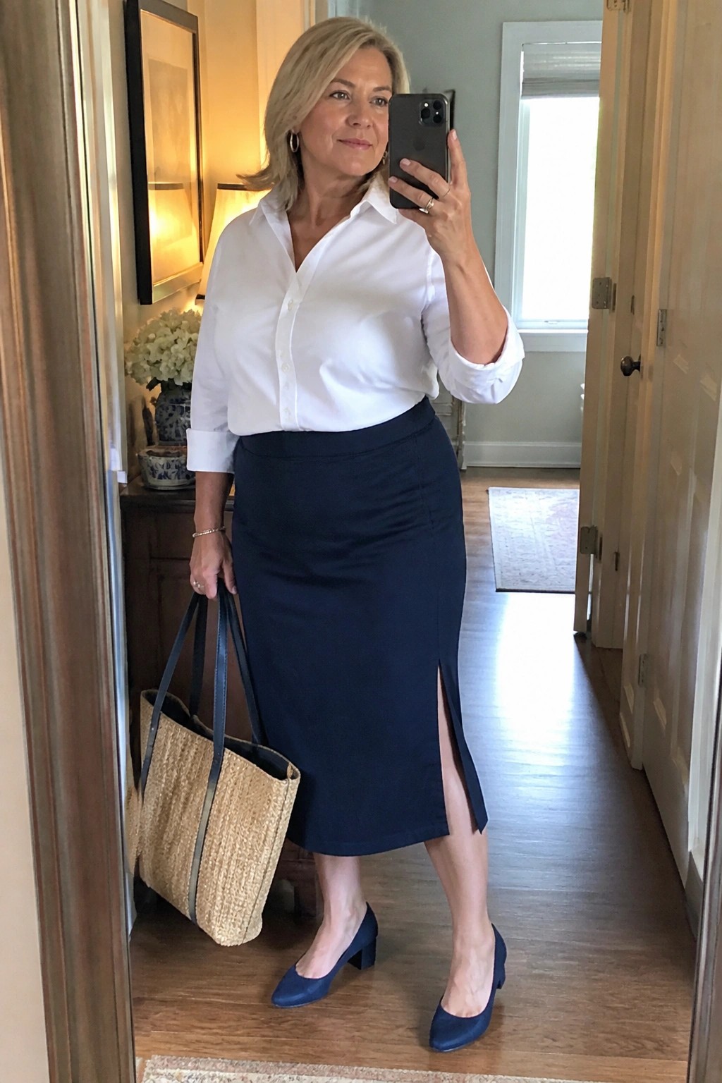 Woman in white shirt and navy skirt holding phone and woven tote in hallway