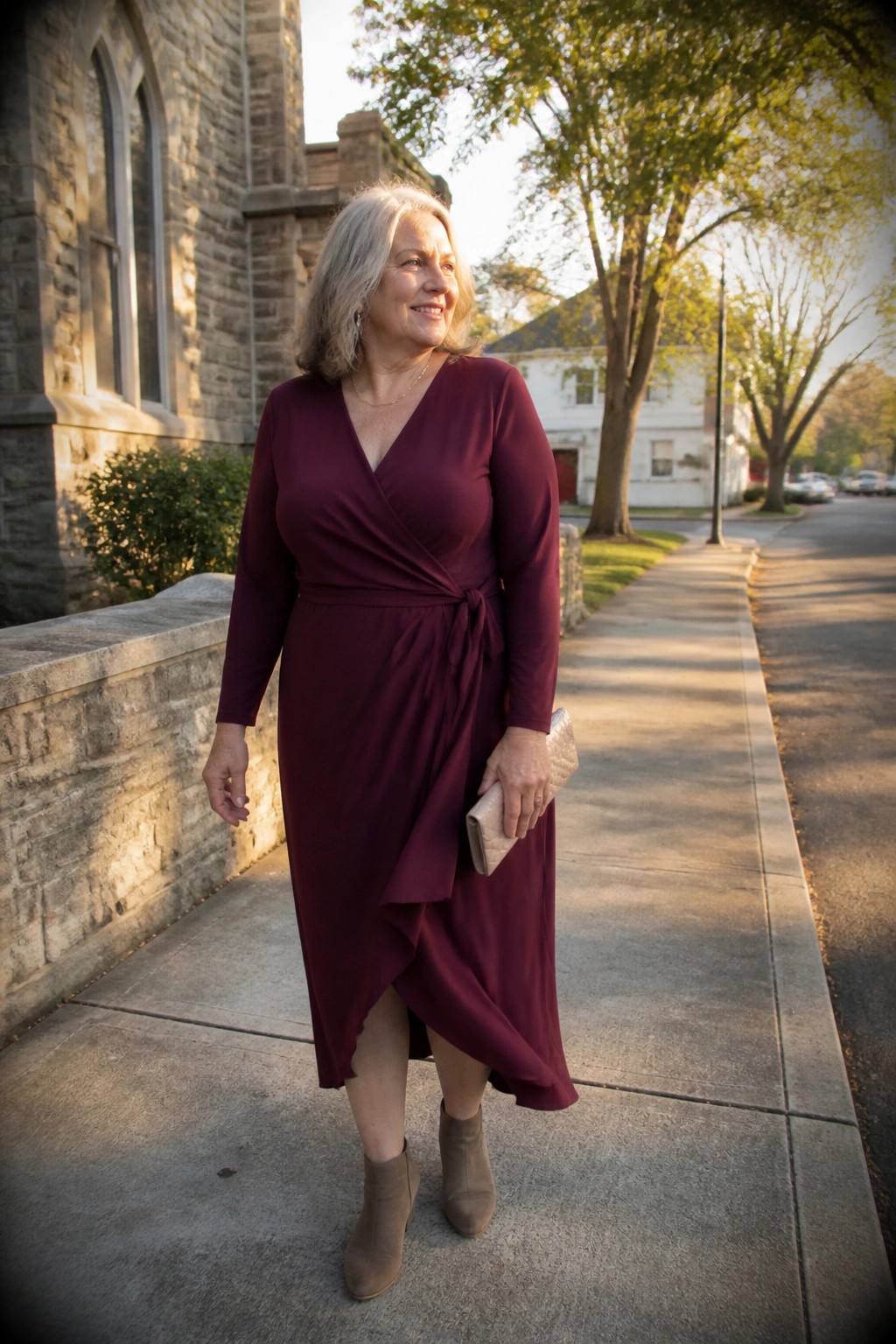 Gray-haired woman in maroon wrap dress holding clutch on sidewalk by stone wall