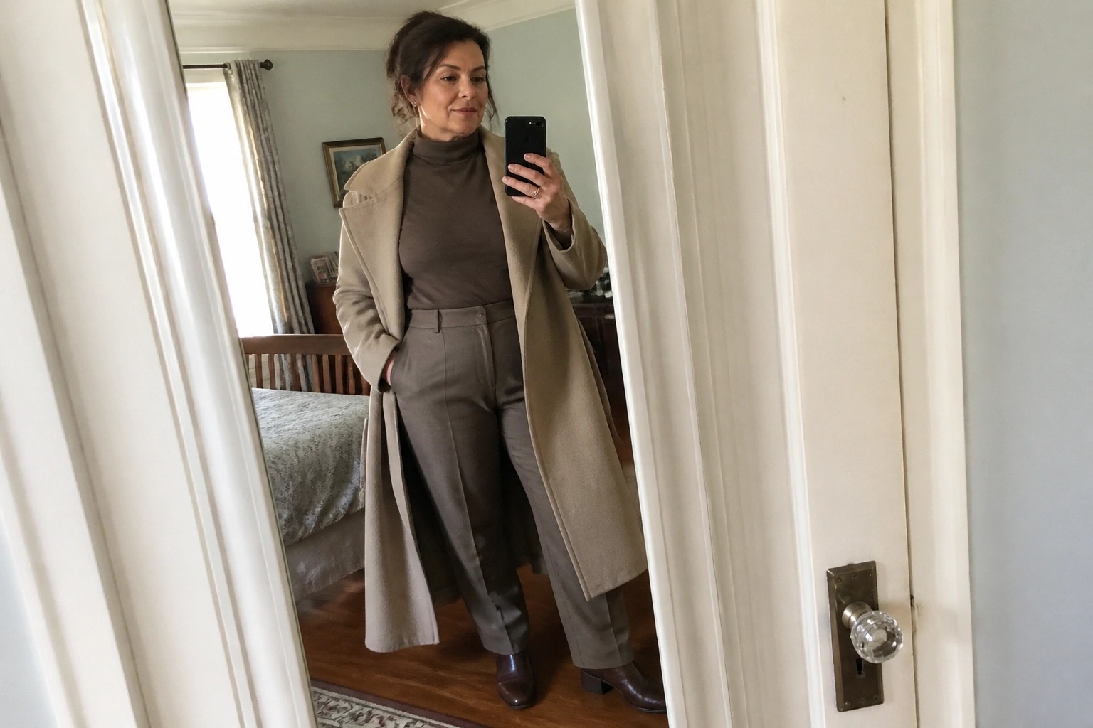 Woman in beige coat and brown pants taking mirror selfie in bedroom