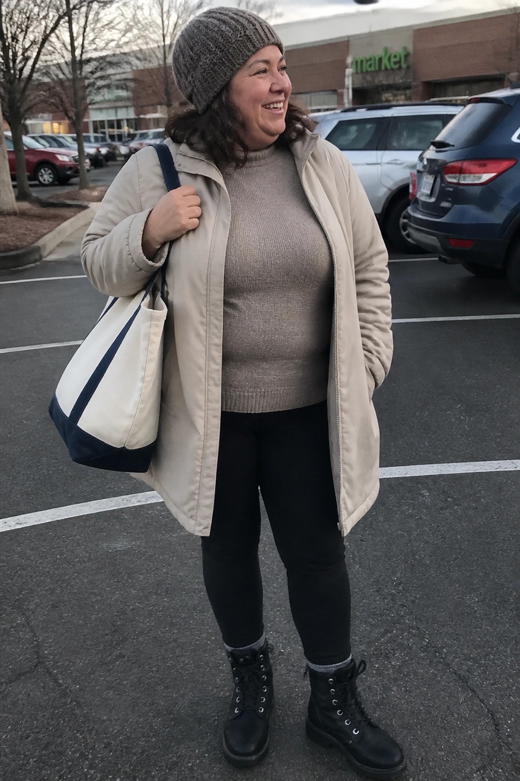 Woman in gray beanie and beige jacket smiles in parking lot with tote bag