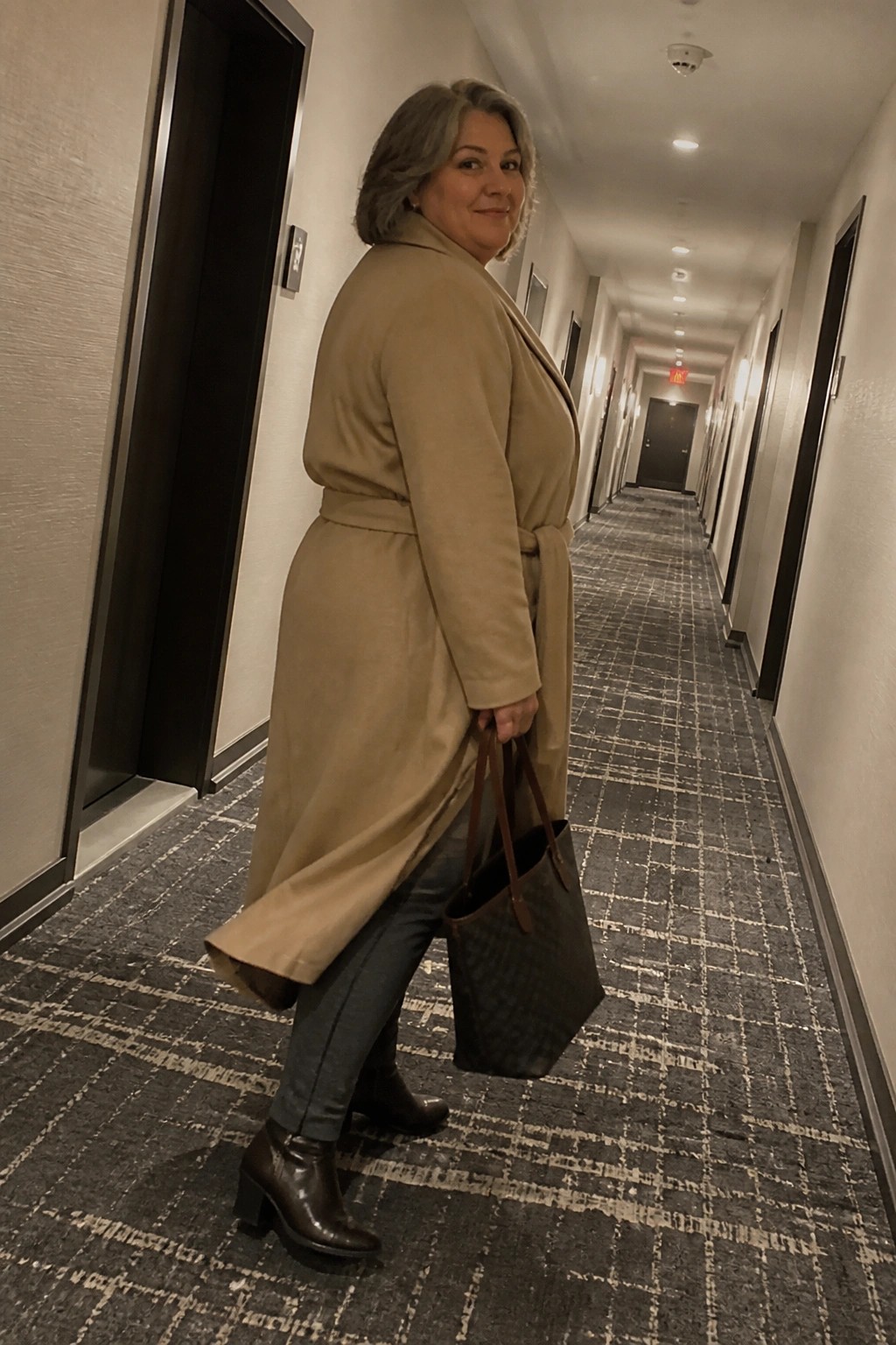 Woman in beige coat carrying tote bag in hotel hallway