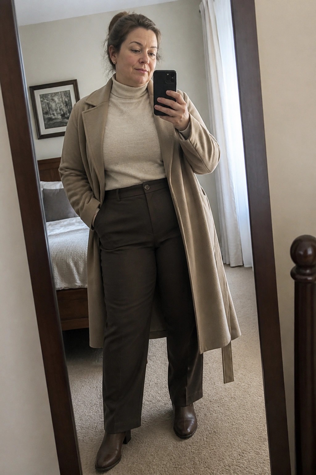 Woman in beige coat and brown pants taking mirror selfie in bedroom