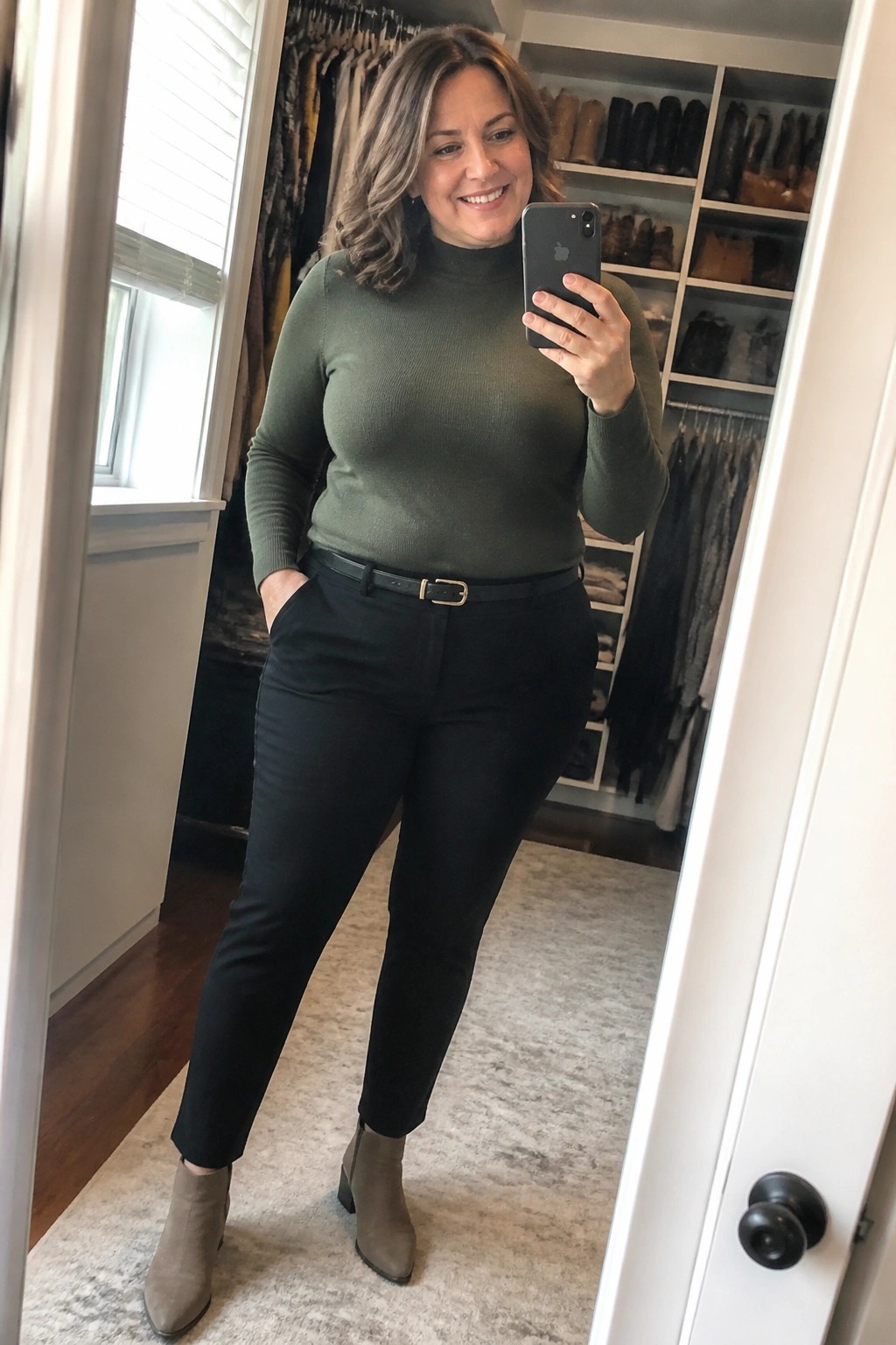 Woman in green turtleneck and black pants taking mirror selfie in closet