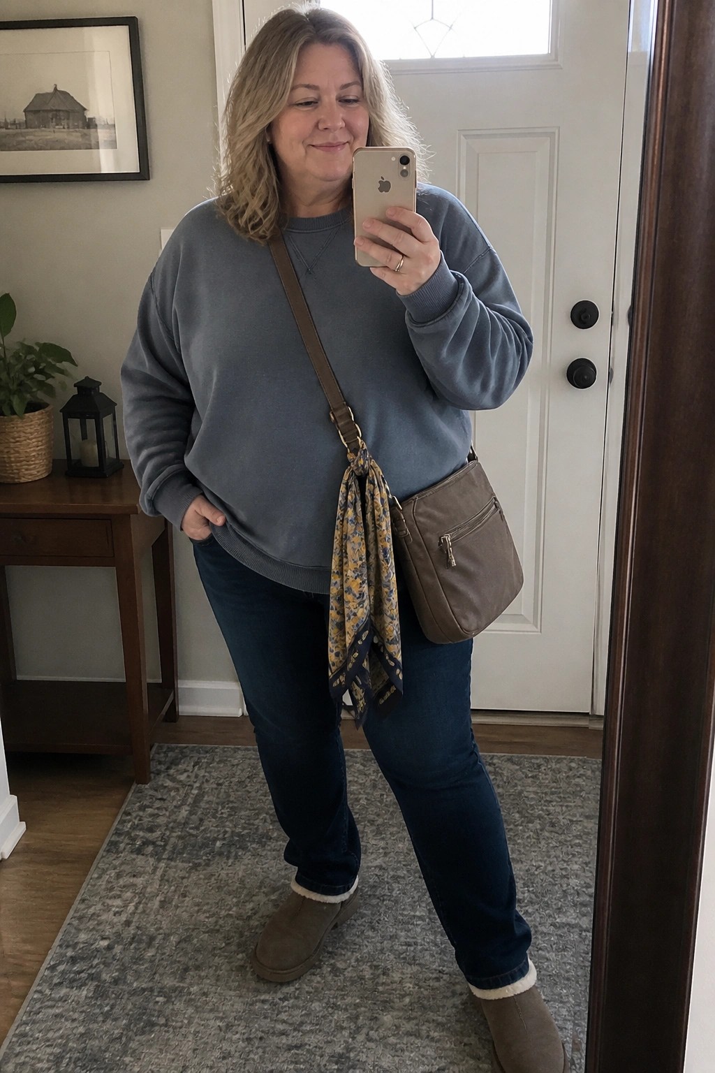 Woman in blue sweatshirt taking mirror selfie with floral scarf on crossbody bag